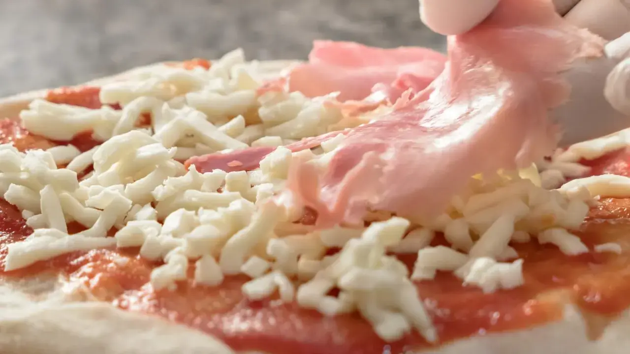 Rękawiczki zakładają cienkie plastry szynki parmeńskiej na pizzę z serem i sosem pomidorowym.