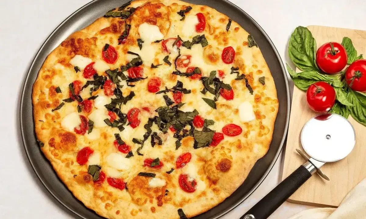 Pyszna pizza z serka wiejskiego, pomidorkami koktajlowymi i bazylią. Obok leży nóż do pizzy i świeże pomidory.