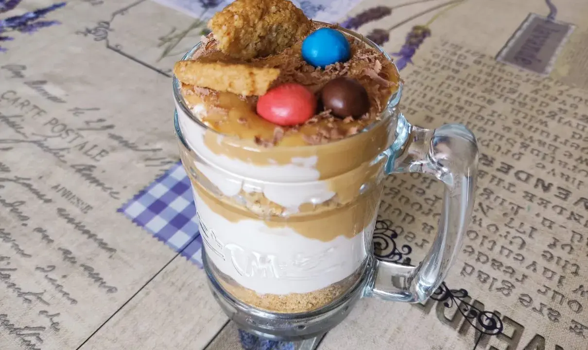 Warstwowy deser banoffee w szklance z polewą karmelową, bitą śmietaną, ciasteczkami i kolorowymi drażetkami.