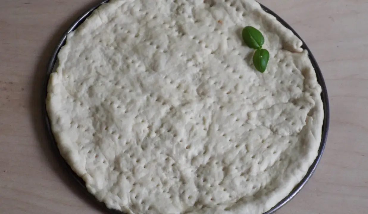 Gotowy do pieczenia, okrągły spód do pizzy z dziurkami, ozdobiony listkiem bazylii. Idealny szybki przepis na ciasto do pizzy.