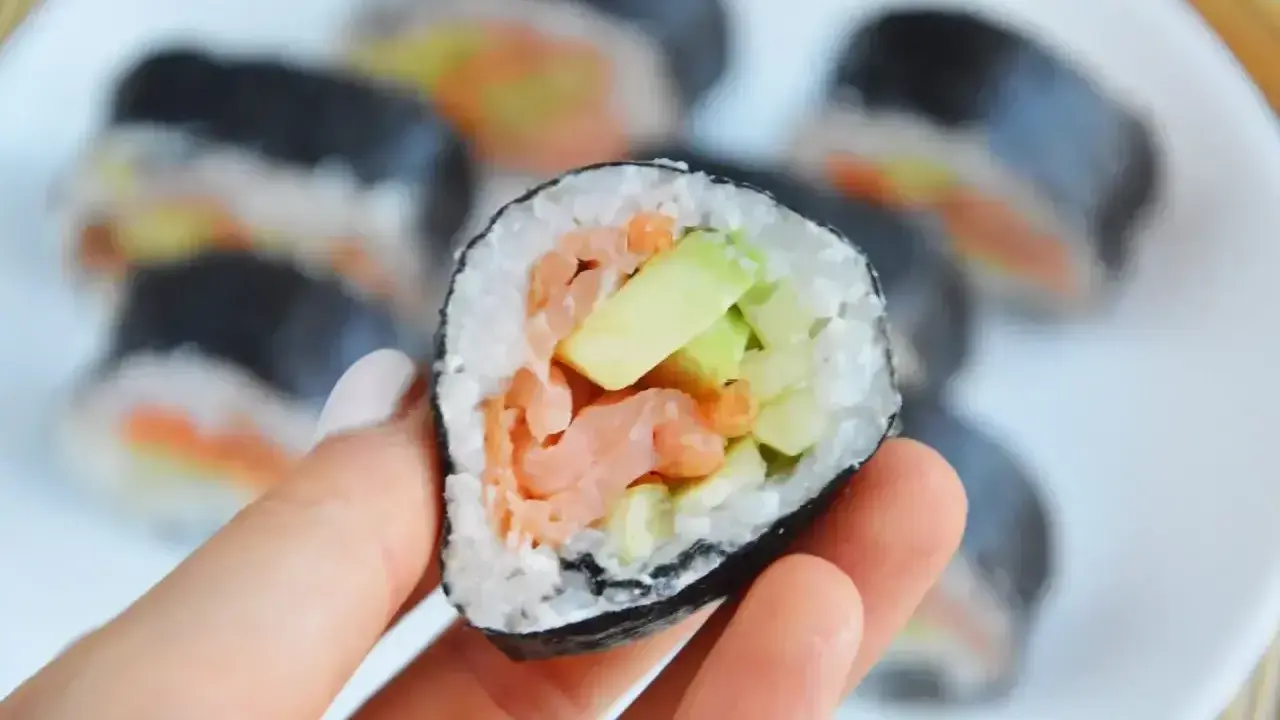 Ręka trzyma kawałek futomaka z łososiem i awokado. Na talerzu widać więcej sushi.
