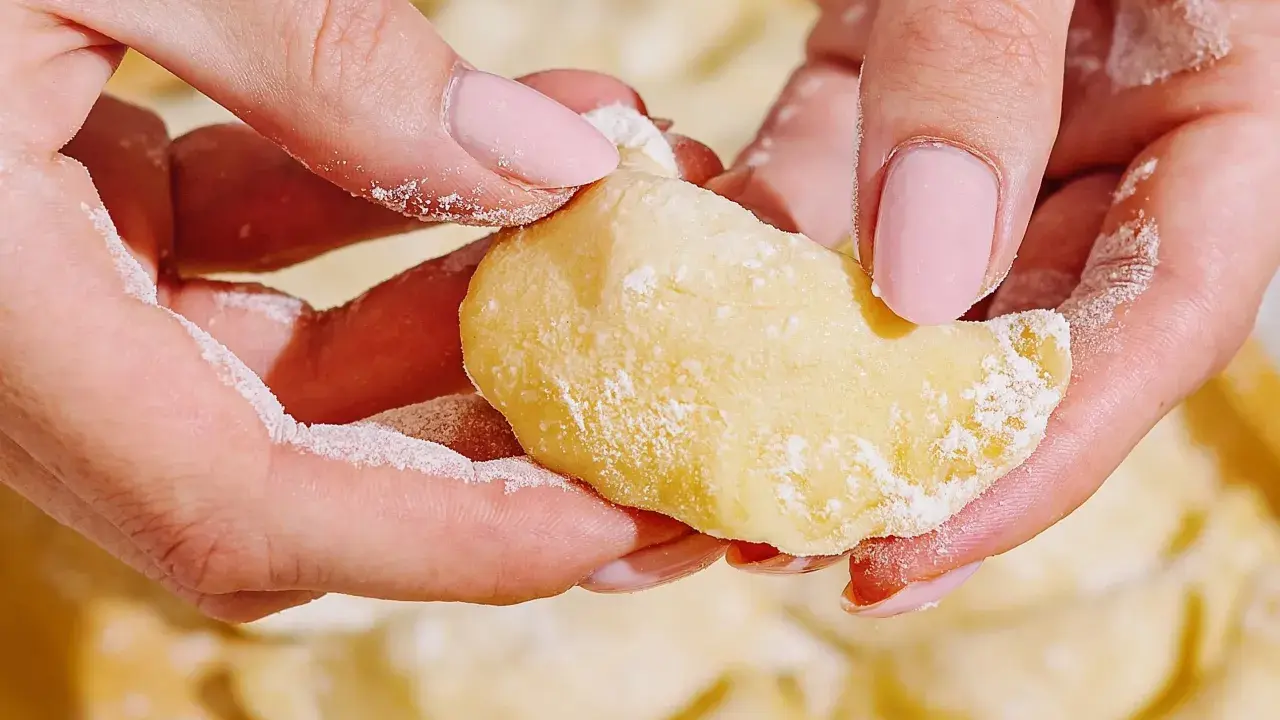 Dłonie z różowymi paznokciami lepią pierogi. Mąka lekko oprósza palce i ciasto. To idealny przepis na domowe pierogi.