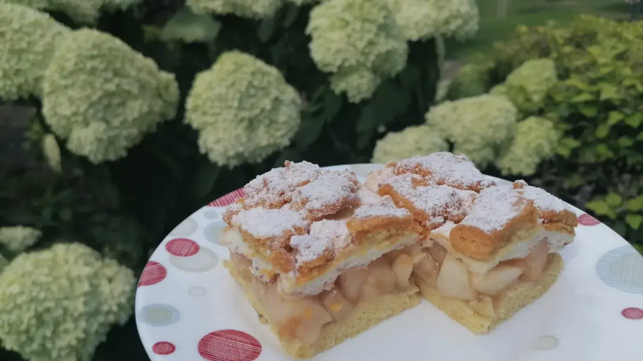 Pyszna szarlotka z bezą i kruszonką, oprószona cukrem pudrem, na talerzu wśród kwitnących hortensji.