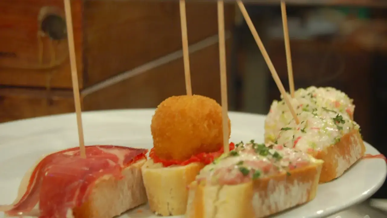 Różnorodne pinchos na białym talerzu: z szynką, kulką w panierce, pastą rybną i sałatką.