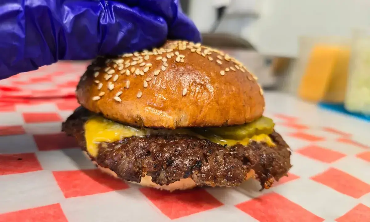Rękawiczka w niebieskiej rękawiczce przygotowuje pysznego smash burgera z serem i ogórkiem.