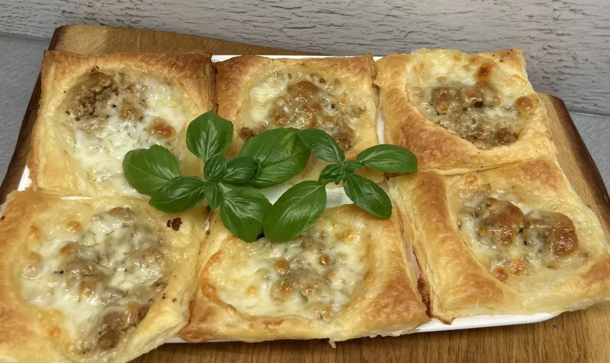 Sześć kwadratowych porcji ciasta francuskiego z mięsem mielonym i serem, ozdobione listkami bazylii.
