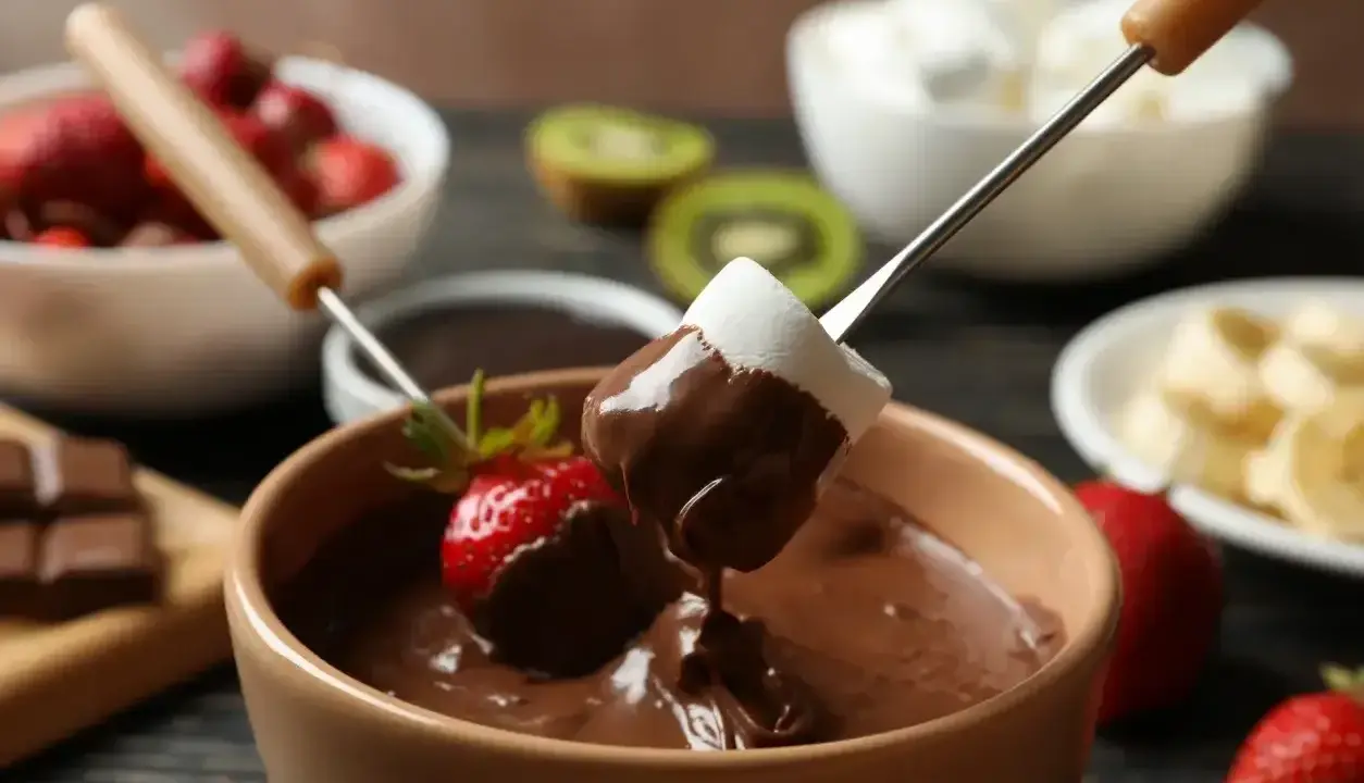 Pyszne fondue czekoladowe z pianką i truskawką na patyczku. W tle owoce i pianki.