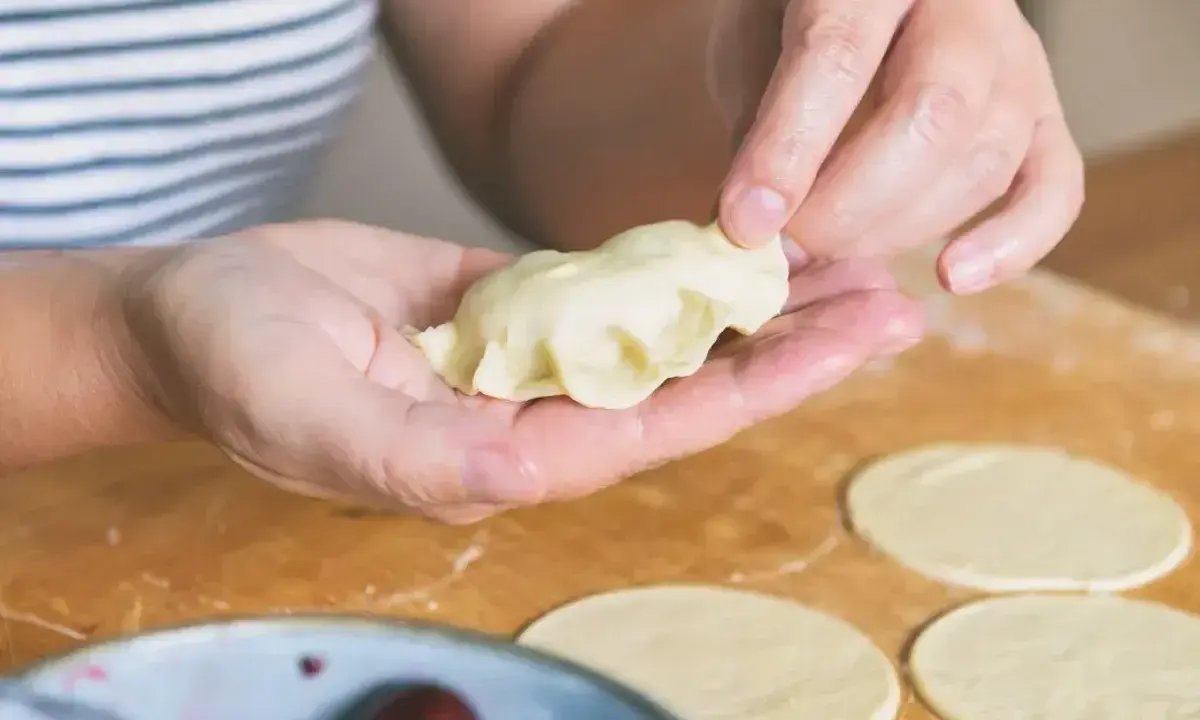 Dłonie formują pieroga z ciasta na pierogi bez jajka. Na blacie leżą okrągłe placki ciasta i miska z wiśniami.