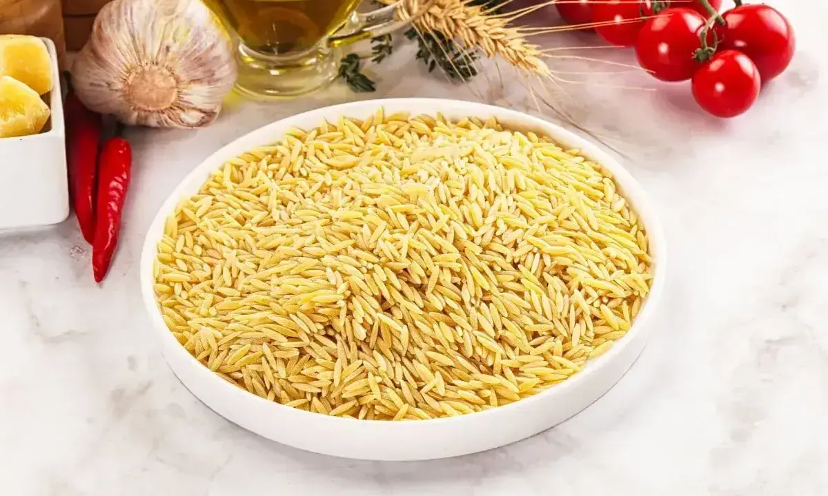 Biała miska pełna makaronu w kształcie ryżu, obok czosnek, chili, oliwa i pomidorki.