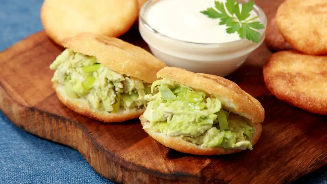 Dwie pyszne arepas z kurczakiem i awokado, obok miseczka sosu i kilka niefaszerowanych arepas.