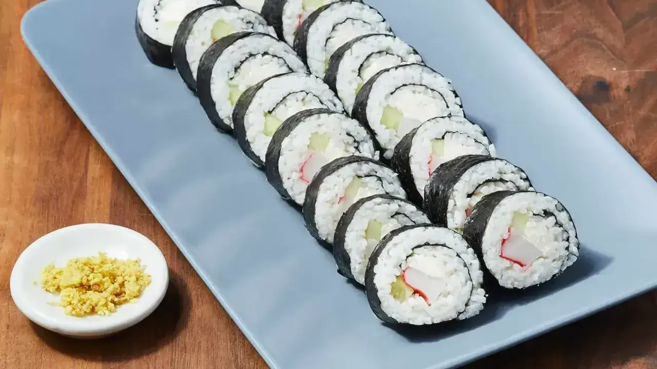 Rząd roladek sushi z kremowym serkiem, ogórkiem i paluszkami krabowymi na szarym talerzu. Obok mała miseczka z imbirem.