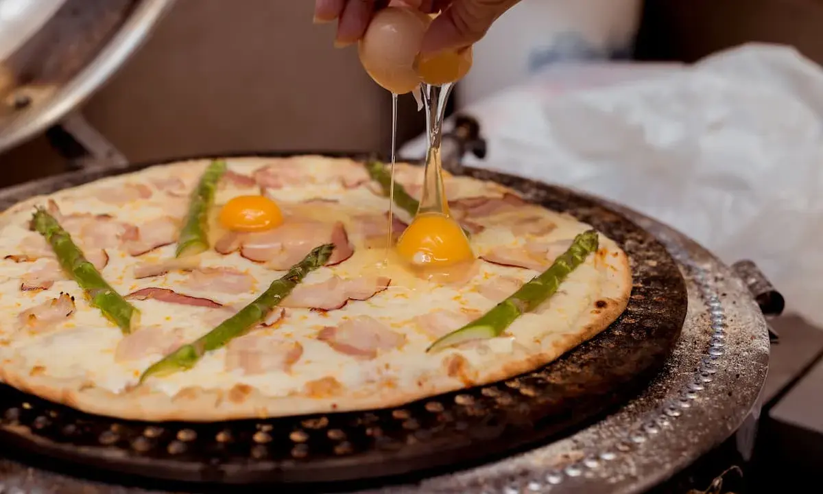 Kucharz dodaje jajko do pizzy z boczkiem i szparagami. Pyszna pizza z jajkiem w przygotowaniu.