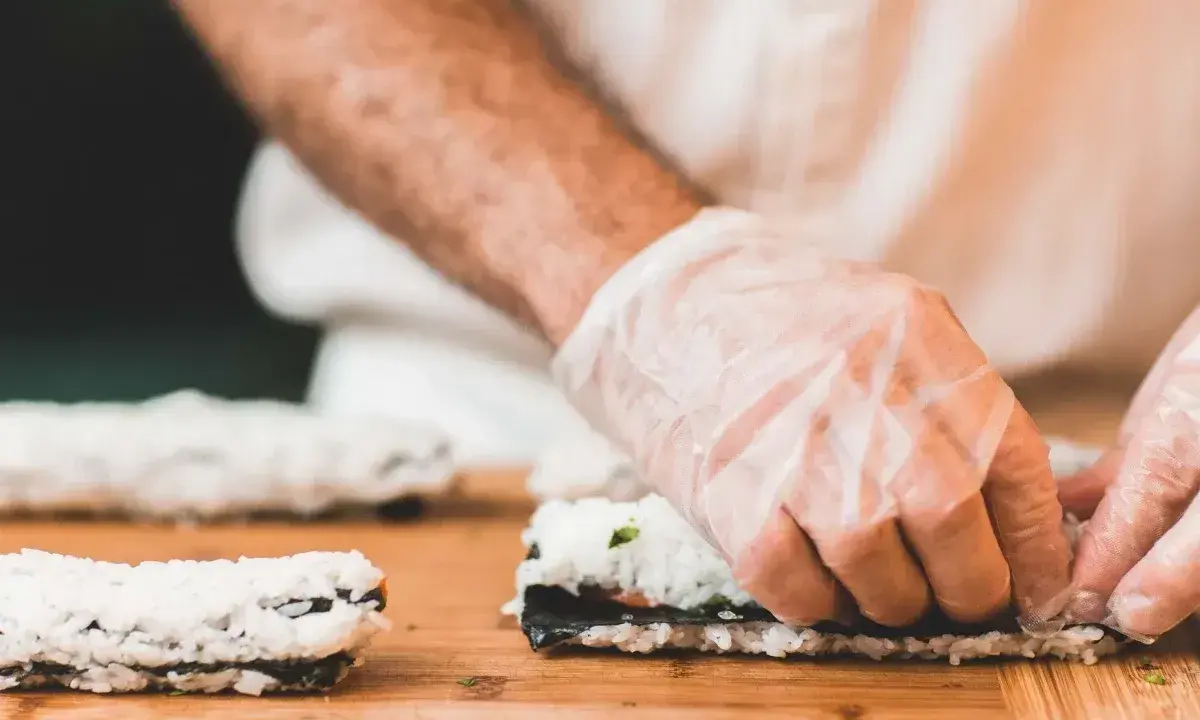 Ręce w rękawiczkach zwijają sushi. Na desce leżą gotowe rolki i składniki do kolejnej. Idealna zalewa do sushi podkreśli smak.