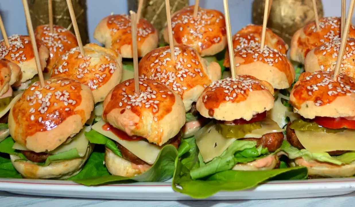 Mini hamburgery z sezamem na patyczkach, ułożone na talerzu z sałatą i dodatkami.