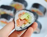 Ręka trzyma kawałek futomaka z łososiem i awokado. Na talerzu widać więcej sushi.