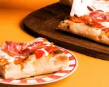 Kawałek pysznej pizzy rzymskiej z szynką i ciągnącym się serem na talerzu, obok reszta na desce.
