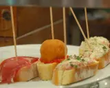 Różnorodne pinchos na białym talerzu: z szynką, kulką w panierce, pastą rybną i sałatką.