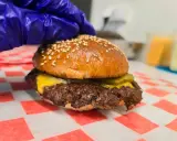 Rękawiczka w niebieskiej rękawiczce przygotowuje pysznego smash burgera z serem i ogórkiem.