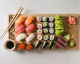 Deska z różnorodnymi składnikami do sushi: nigiri z łososiem, tuńczykiem i krewetkami, maki z awokado i rybą, rolki w ikrze, sashimi z tuńczyka, sos sojowy, wasabi i marynowany imbir.