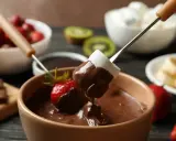 Pyszne fondue czekoladowe z pianką i truskawką na patyczku. W tle owoce i pianki.