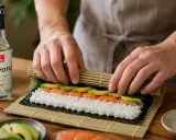 Ręce zwijają domowe sushi z łososiem i awokado na macie bambusowej. Obok butelka octu ryżowego.