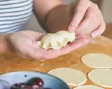 Dłonie formują pieroga z ciasta na pierogi bez jajka. Na blacie leżą okrągłe placki ciasta i miska z wiśniami.