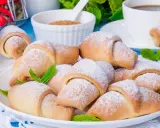 Kruche rogaliki z marmoladą, posypane cukrem pudrem, ozdobione listkami mięty, gotowe do podania z kawą.