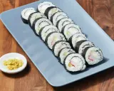Rząd roladek sushi z kremowym serkiem, ogórkiem i paluszkami krabowymi na szarym talerzu. Obok mała miseczka z imbirem.