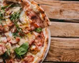 Pyszna pizza carbonara z chrupiącym boczkiem, rukolą i cebulą na drewnianym stole.