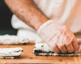Ręce w rękawiczkach zwijają sushi. Na desce leżą gotowe rolki i składniki do kolejnej. Idealna zalewa do sushi podkreśli smak.