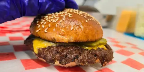 Rękawiczka w niebieskiej rękawiczce przygotowuje pysznego smash burgera z serem i ogórkiem.
