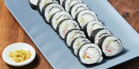 Rząd roladek sushi z kremowym serkiem, ogórkiem i paluszkami krabowymi na szarym talerzu. Obok mała miseczka z imbirem.
