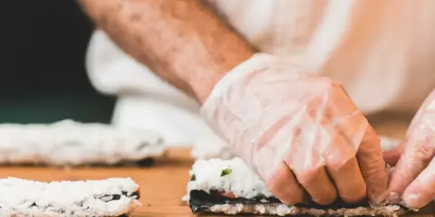 Ręce w rękawiczkach zwijają sushi. Na desce leżą gotowe rolki i składniki do kolejnej. Idealna zalewa do sushi podkreśli smak.