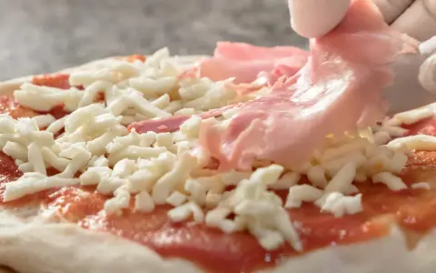 Rękawiczki zakładają cienkie plastry szynki parmeńskiej na pizzę z serem i sosem pomidorowym.