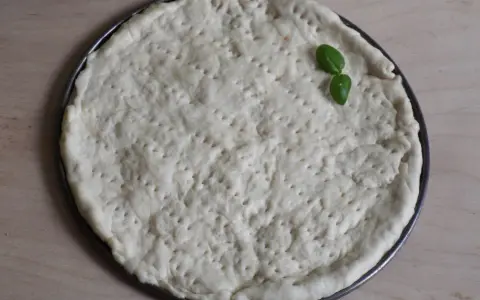 Gotowy do pieczenia, okrągły spód do pizzy z dziurkami, ozdobiony listkiem bazylii. Idealny szybki przepis na ciasto do pizzy.