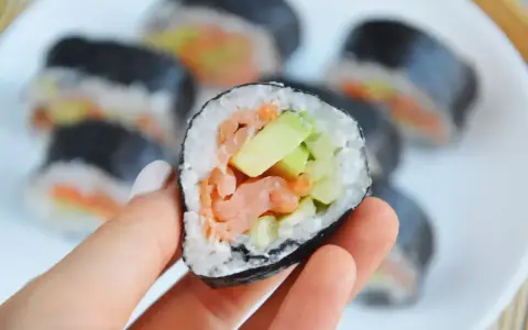 Ręka trzyma kawałek futomaka z łososiem i awokado. Na talerzu widać więcej sushi.