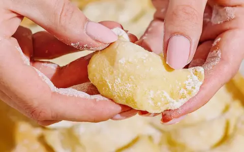 Dłonie z różowymi paznokciami lepią pierogi. Mąka lekko oprósza palce i ciasto. To idealny przepis na domowe pierogi.
