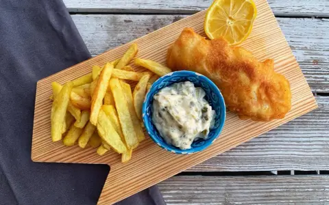 Chrupiące fish and chips z cytryną i sosem tatarskim na drewnianej desce.