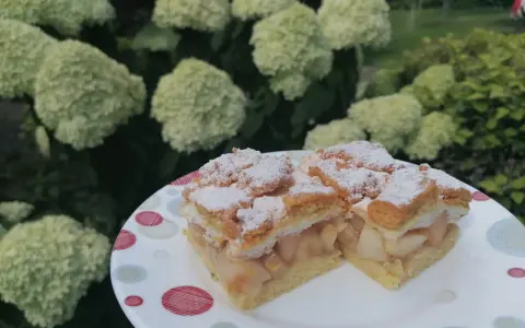Pyszna szarlotka z bezą i kruszonką, oprószona cukrem pudrem, na talerzu wśród kwitnących hortensji.