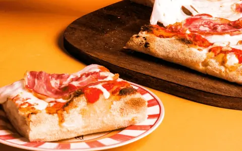 Kawałek pysznej pizzy rzymskiej z szynką i ciągnącym się serem na talerzu, obok reszta na desce.