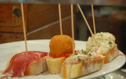 Różnorodne pinchos na białym talerzu: z szynką, kulką w panierce, pastą rybną i sałatką.