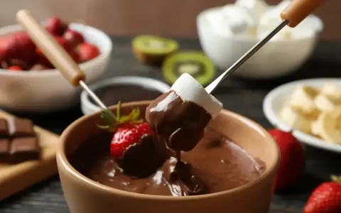 Pyszne fondue czekoladowe z pianką i truskawką na patyczku. W tle owoce i pianki.