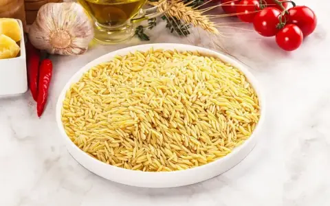 Biała miska pełna makaronu w kształcie ryżu, obok czosnek, chili, oliwa i pomidorki.