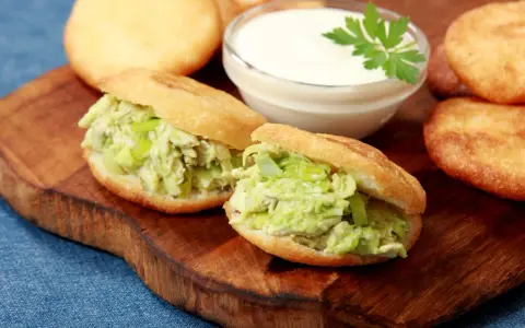 Dwie pyszne arepas z kurczakiem i awokado, obok miseczka sosu i kilka niefaszerowanych arepas.