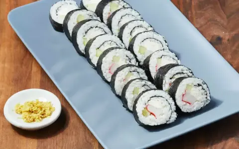 Rząd roladek sushi z kremowym serkiem, ogórkiem i paluszkami krabowymi na szarym talerzu. Obok mała miseczka z imbirem.