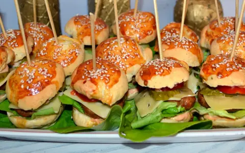 Mini hamburgery z sezamem na patyczkach, ułożone na talerzu z sałatą i dodatkami.
