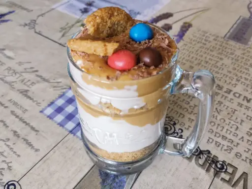 Warstwowy deser banoffee w szklance z polewą karmelową, bitą śmietaną, ciasteczkami i kolorowymi drażetkami.