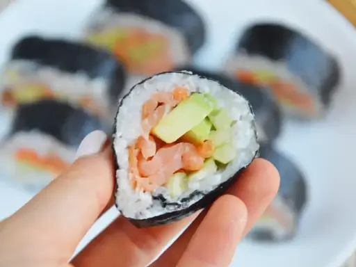 Ręka trzyma kawałek futomaka z łososiem i awokado. Na talerzu widać więcej sushi.
