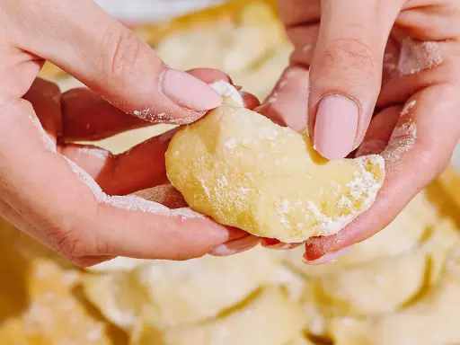 Dłonie z różowymi paznokciami lepią pierogi. Mąka lekko oprósza palce i ciasto. To idealny przepis na domowe pierogi.