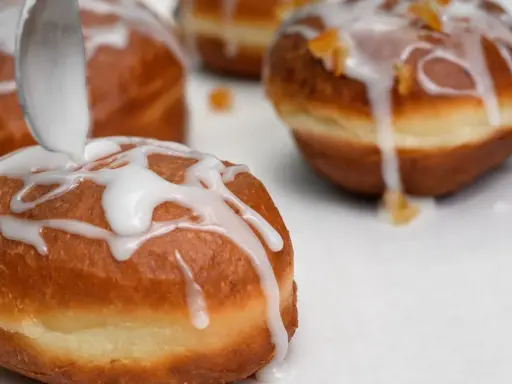 Lukier przepis na pączki. Łyżka polewa słodką masą pączki, które zaraz trafią na stół.